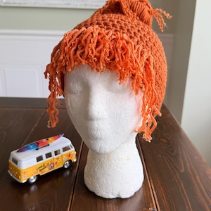 May include: A bright orange crocheted hat featuring a textured, knotted top and long, curly fringe detailing that hangs around the forehead and sides. The hat is displayed on a white mannequin head, positioned next to a small yellow toy van.