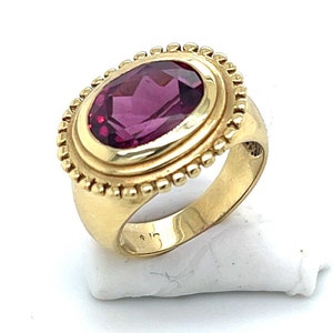 May include: A gold ring with a large oval-shaped purple gemstone set in a bezel setting. The ring has a textured band and a decorative border around the gemstone.