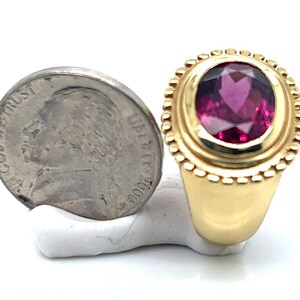May include: A gold ring with a large oval-shaped pink gemstone set in a bezel setting. The ring is surrounded by a decorative border of small gold beads.