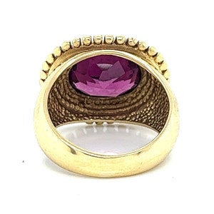 May include: A gold ring with a large, oval-shaped, purple gemstone set in a bezel setting. The ring has a textured band with a beaded edge.