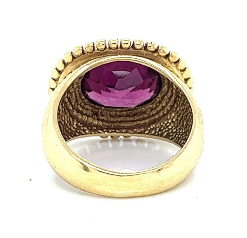 May include: A gold ring with a large, oval-shaped, purple gemstone set in a bezel setting. The ring has a textured band with a beaded edge.