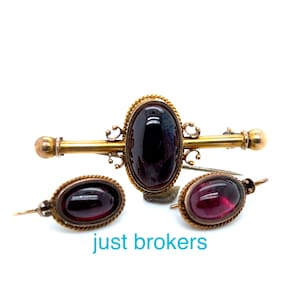 May include: A set of three antique gold-tone brooches with red glass cabochons. The brooches are in the shape of ovals and have a rope-like border. The largest brooch is a bar pin with a decorative scroll design.
