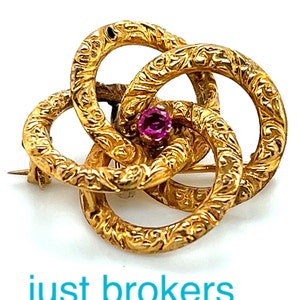 May include: A gold brooch with a pink stone in the centre. The brooch is made of three interlocking circles with a textured surface.
