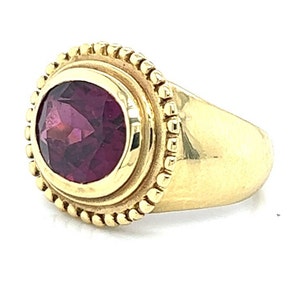 May include: A gold ring with a deep purple oval gemstone set in a bezel setting. The ring has a beaded edge around the bezel.