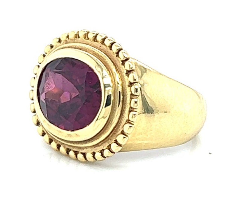 May include: A gold ring with a deep purple oval gemstone set in a bezel setting. The ring has a beaded edge around the bezel.