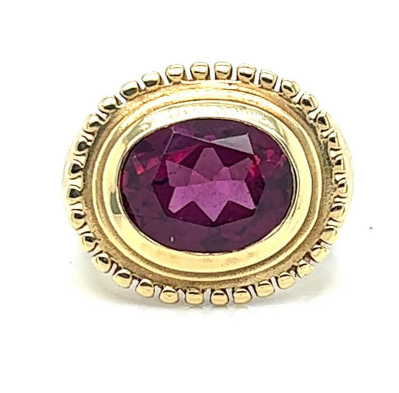 May include: A gold ring with a deep red oval gemstone set in a bezel setting. The ring has a decorative border of small beads.