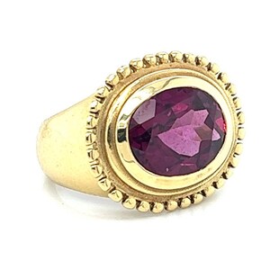 May include: A gold ring with a deep purple oval gemstone set in a bezel setting. The ring has a beaded edge around the bezel.