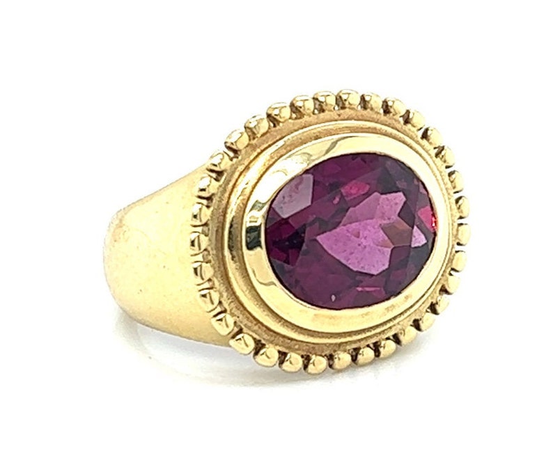 May include: A gold ring with a deep purple oval gemstone set in a bezel setting. The ring has a beaded edge around the bezel.