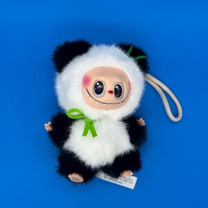 May include: A plush panda-like doll with a smiling face, black and white fur, and a green bow. The doll has a cream-colored face with rosy cheeks and large, black eyes. A white tag is attached to the doll.