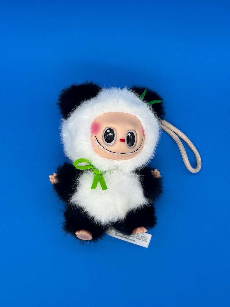 May include: A plush panda-like doll with a smiling face, black and white fur, and a green bow. The doll has a cream-colored face with rosy cheeks and large, black eyes. A white tag is attached to the doll.