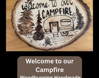 May include: A wood-burned sign with the text "Welcome to our Campfire." The sign features a drawing of a camper, trees, and a campfire. The sign is handmade and has a natural wood edge.