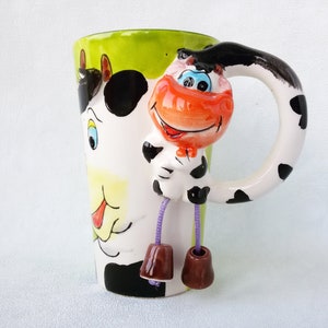 May include: A white ceramic mug with a green, black, and white cow design. The handle is shaped like a cow's tail and has black spots. The cow has a red nose and blue eyes.