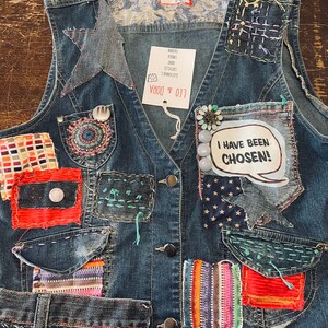 May include: A blue denim vest with various patches and embellishments. The patches include a star, a circle, a square, and a rectangle. The vest has two pockets and two buttons. The text "I HAVE BEEN CHOSEN!" is on one of the patches. The vest has a tag that says "LEO & DORA".