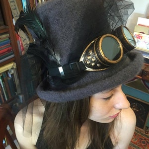May include: A gray felt top hat with a black veil and gold-toned goggles. The hat has a black band with a silver buckle.