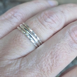 May include: Three silver rings with a textured, twisted design are stacked on a hand. The rings are thin and appear to be made of metal. The hand is in soft focus, with the rings as the focal point.