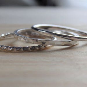 May include: Three silver rings with varying textures are displayed on a light wooden surface. One ring features a twisted rope design, another has a hammered texture, and the third is smooth. These rings are suitable for everyday wear.