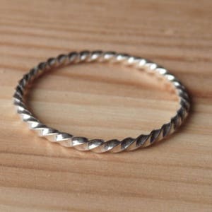 Peut inclure: Une bague argentée avec un motif torsadé en forme de corde. La bague est circulaire et repose sur une surface en bois brun clair. La texture de la bague est visible, chaque torsion reflétant la lumière.