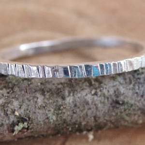 May include: A silver ring with a textured surface. The ring is set against a natural, wooden background. The ring's surface has a series of vertical lines, giving it a unique, handcrafted appearance. This ring is a piece of jewellery.
