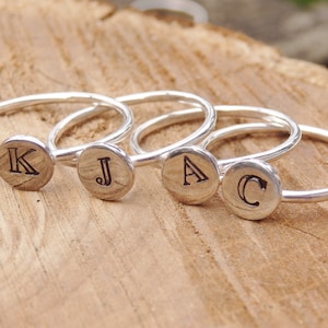 May include: Four silver rings, each with a circular wooden disc bearing the letters K, J, A, and C. The rings are set against a wooden background, highlighting the engraved initials. The rings have a delicate, minimalist design.