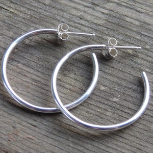 May include: A pair of silver hoop earrings with post backs. The earrings are round and have a polished finish. The earrings are displayed on a weathered wooden surface.