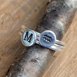 Peut inclure: Trois bagues en argent empilées. La bague du haut porte la lettre "M" gravée. La bague du milieu porte un motif de vague stylisé gravé. La bague du bas est unie.