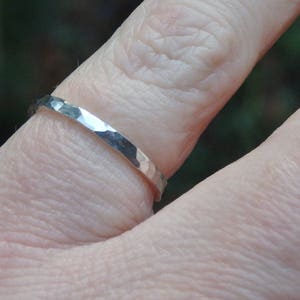 May include: A silver ring with a hammered texture is worn on a finger. The ring has a simple band design and reflects light, showcasing its metallic sheen. The background is blurred, emphasising the ring's details.
