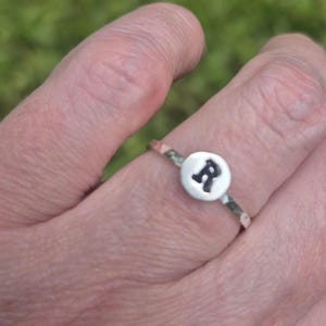 Peut inclure: Une bague argentée dotée d'un anneau fin martelé et d'un petit disque blanc circulaire en guise de pièce centrale. Le disque est orné d'une lettre R majuscule noire en relief, écrite dans une police à empattement, contrastant avec le blanc.