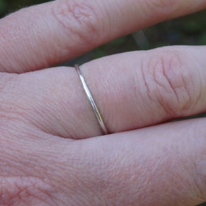 May include: A slender, silver ring is worn on a finger. The ring has a simple, minimalist design. The ring is approximately 1 mm wide. The skin tone of the person wearing the ring is fair.
