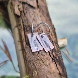 May include: Copper-toned rectangular earrings with a botanical design. Each earring has a small, etched tree design and a silver-coloured hook. The earrings are displayed on a piece of wood.