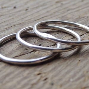 May include: Three silver rings are interlocked, set against a weathered wooden surface. The rings are slender and have a polished, reflective finish. The rings are likely made of sterling silver or a similar metal. The rings are a simple, elegant jewellery item.