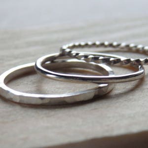 May include: A set of three silver rings stacked together. The first ring has a flat, hammered texture. The second ring is a smooth, plain band. The third ring features a twisted, rope-like design. All three rings are metallic silver in colour.