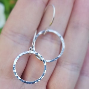 May include: Silver hoop earrings with a hammered finish. The earrings have a simple, minimalist design, featuring a circular shape with a hook closure. The earrings are shown against a blurred background.