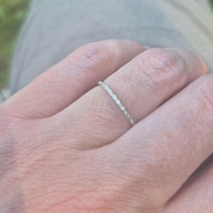 May include: A thin, silver-coloured ring with a textured, uneven surface. The band features a metallic finish with small, irregular indentations along its circumference, creating a rugged appearance on the slender, circular metal jewellery piece.