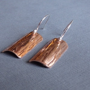 May include: A pair of copper earrings with a hammered texture and silver hooks. The earrings are rectangular in shape and have a slightly curved edge.