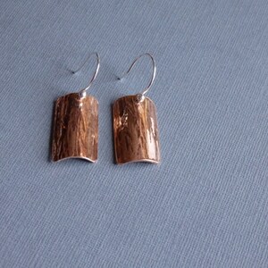 May include: A pair of silver earrings with a hammered copper rectangular pendant. The earrings have a simple wire hook closure.