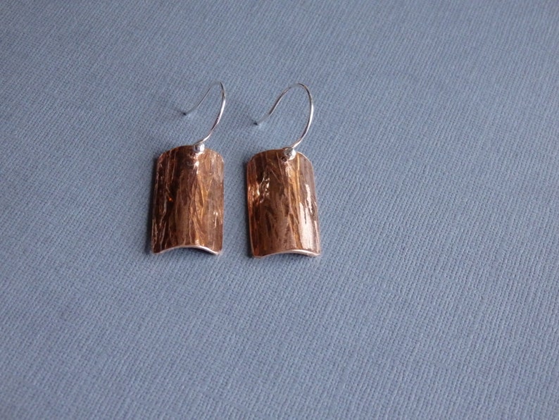 May include: A pair of silver earrings with a hammered copper rectangular pendant. The earrings have a simple wire hook closure.