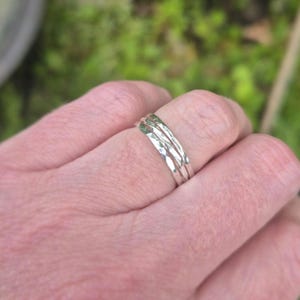 May include: Three silver hammered rings stacked on a finger. The rings have a textured, irregular surface, reflecting light. The background is blurred, with green and brown tones suggesting foliage. The rings are a simple, elegant piece of jewellery.
