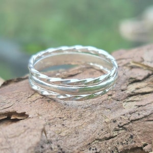 May include: Three silver rings, one with a twisted design, stacked together. The rings are displayed on a piece of brown bark, with a blurred green background.
