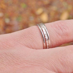 May include: Two silver rings stacked on a finger. Both rings are thin and have a textured surface.