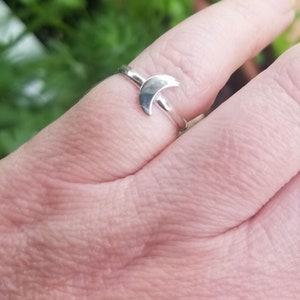 May include: A silver ring with a crescent moon design. The ring is worn on a finger, with the moon shape clearly visible. The ring's band is slender and plain, enhancing the moon's graceful curve. The background is blurred, with green leaves in view.