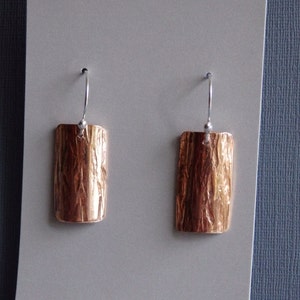 May include: A pair of copper earrings with a textured, wood-like finish. The earrings are rectangular in shape and have a silver hook.