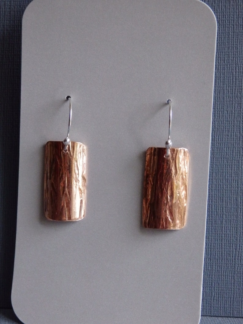 May include: A pair of copper earrings with a textured, wood-like finish. The earrings are rectangular in shape and have a silver hook.