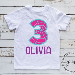 Puede incluir: Camiseta blanca con un n&uacute;mero 3 rosa con un dise&ntilde;o de telara&ntilde;a azul. El nombre Olivia est&aacute; escrito en morado debajo del n&uacute;mero.