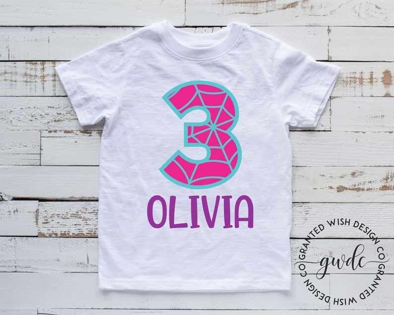 Puede incluir: Camiseta blanca con un n&uacute;mero 3 rosa con un dise&ntilde;o de telara&ntilde;a azul. El nombre Olivia est&aacute; escrito en morado debajo del n&uacute;mero.