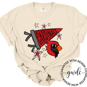 May include: Cream-colored t-shirt featuring a graphic of a red cardinal bird and a pennant that reads "CARDINALS" in red letters. The design includes star accents in red and white. The shirt is knotted at the waist.