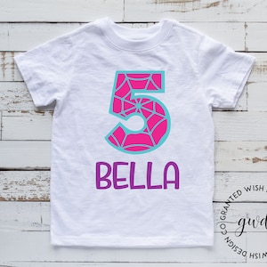 Puede incluir: Camiseta blanca con un n&uacute;mero 5 rosa y azul con un dise&ntilde;o de telara&ntilde;a. El nombre "Bella" est&aacute; escrito en morado debajo del n&uacute;mero.