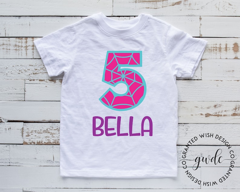 Puede incluir: Camiseta blanca con un n&uacute;mero 5 rosa y azul con un dise&ntilde;o de telara&ntilde;a. El nombre "Bella" est&aacute; escrito en morado debajo del n&uacute;mero.