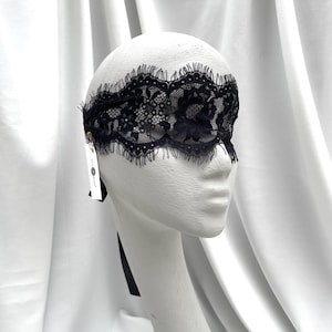 May include: Black lace eye mask with a delicate floral pattern, displayed on a white mannequin head. The mask has a scalloped edge and a black ribbon tie. A small tag hangs from the side.