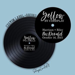 Op de afbeelding: Een zwarte vinylplaat met de tekst "Yellow by Coldplay" en gepersonaliseerde details. De plaat bevat de namen "Harrison + Riley McDonald" en de datum "October 16, 2021" in witte letters. De achtergrond is lichtblauw.