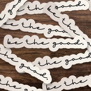 May include: White stickers with black text that reads "be the good." The stickers are arranged in a random pattern on a brown wooden surface.